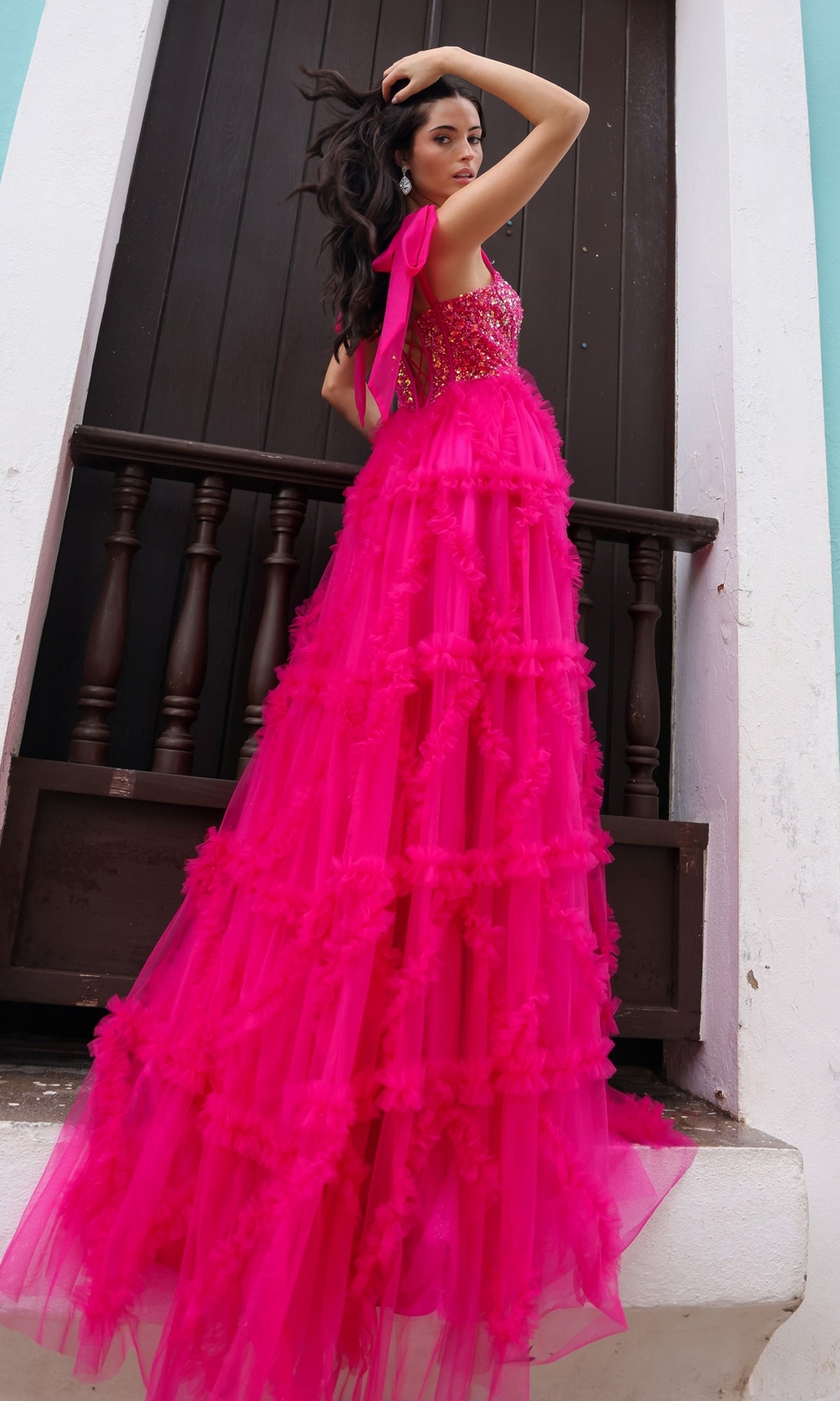 A woman in the Shoulder-Bows Long Ruffled Prom Gown T1340, featuring a bright pink tulle skirt and beaded corset bodice, stands before a dark wooden door, glancing over her shoulder.